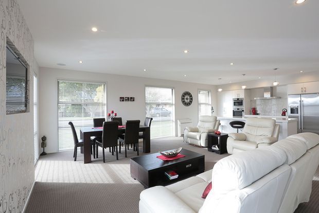 Bright open-plan interior combining a living room, dining area, and kitchen. The space features white leather seating, a dark rectangular coffee table, a dark dining table with black chairs, pale carpet, white walls and cabinetry, recessed ceiling lights, and large windows with blinds. The overall appearance is clean, contemporary, and spacious.
