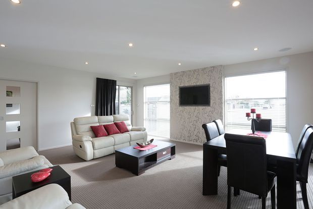 Bright open-plan living and dining area with a cream leather sofa, black coffee table, patterned feature wall with wall-mounted television, and a dark dining table with leather chairs. Large windows with blinds and recessed ceiling lights provide ample natural and artificial light.