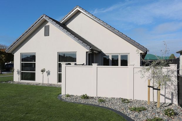 Exterior view of a contemporary single-storey residence with overlapping pitched gables, pale rendered walls, dark-framed windows, and grey tiled roofing. A private courtyard or patio is enclosed by a low pale rendered wall and landscaped with decorative gravel, lawn, and young trees.