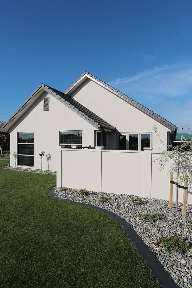Exterior view of a light-coloured single-storey suburban house with overlapping pitched gable roofs, grey tiled roofing, dark-framed windows and doors, and a low rendered courtyard wall. The property is surrounded by a manicured lawn, curved dark edging, and decorative river-stone landscaping.