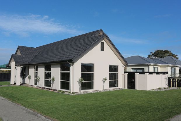 A single-storey suburban house with a light rendered facade, dark tiled gable roof, large black-framed windows, and a neatly landscaped front lawn. A low white boundary fence and neighbouring homes are visible under a clear blue sky.