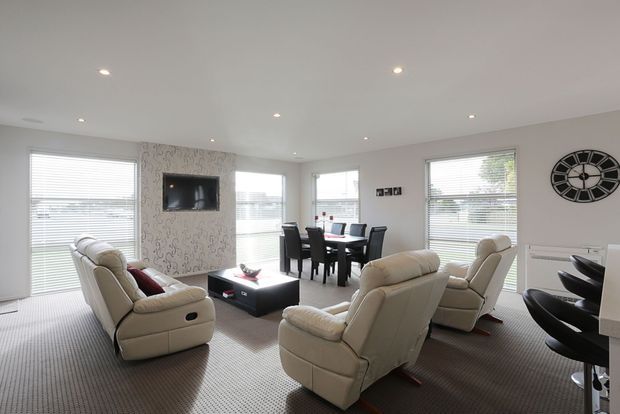 Bright open-plan living and dining area with large windows fitted with white blinds, a patterned feature wall with wall-mounted television, cream reclining leather sofas, a dark coffee table, and a dark dining table with chairs. Recessed ceiling lights and neutral carpeting create a clean, contemporary family interior.