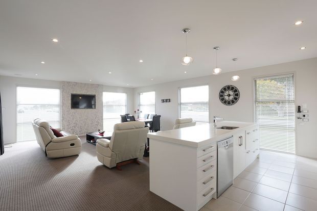 Bright open-plan kitchen and living area with a large white island, integrated dishwasher, pale cabinetry, tiled kitchen floor, carpeted lounge area, upholstered recliner chairs, multiple windows with blinds, pendant lights, recessed ceiling lights, and a wall-mounted clock.