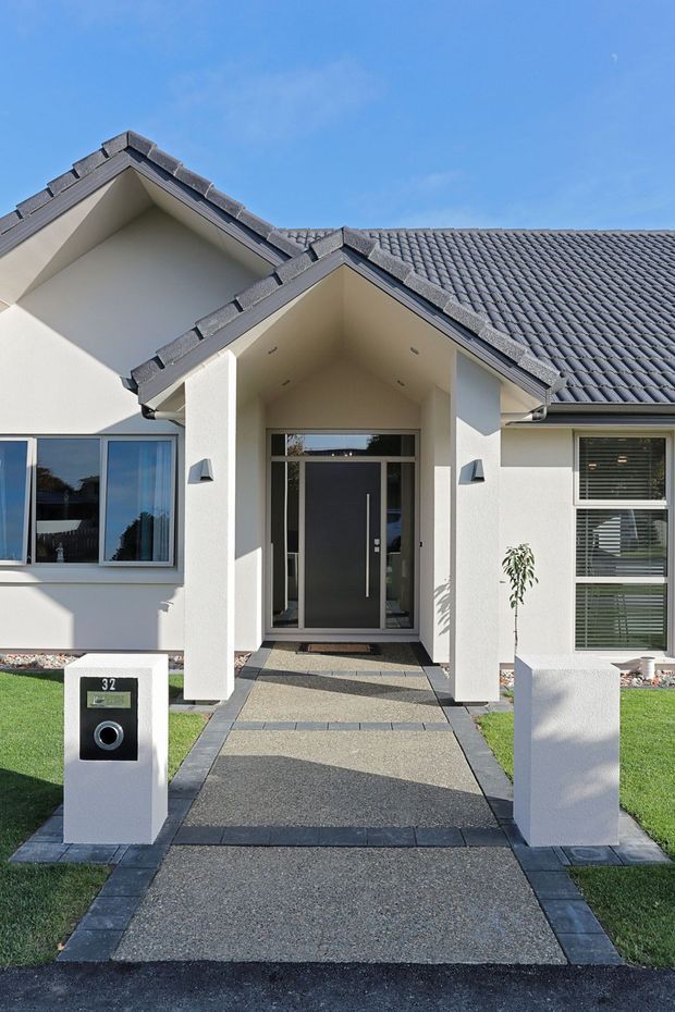 Front exterior view of a contemporary single-storey suburban home with white rendered walls, a dark grey tiled pitched roof, covered entry porch, full-height glazed front door, modern windows, and a formal paved path leading from the street. A minimalist mailbox and low-maintenance lawn frame the entrance.