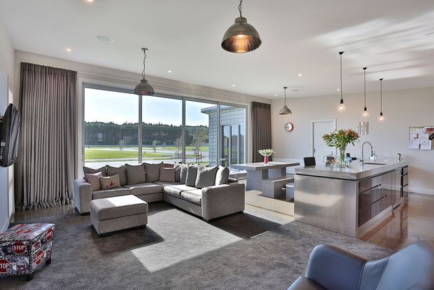 Bright open-plan contemporary living and kitchen space with a large grey sectional sofa, plush carpet, polished timber flooring, pendant lighting, and a stainless-steel kitchen island. Full-height glazing opens to an outdoor area and brings in abundant natural light.