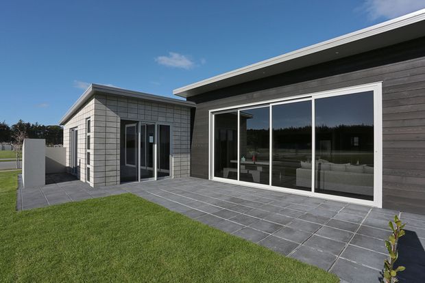 Single-storey contemporary building exterior with a low-pitched roof, dark horizontal timber-look cladding, and a projecting lighter tiled or panelled wing. Large white-framed glazed sliding doors open toward a paved terrace, with a neat lawn and simple boundary wall in the foreground.