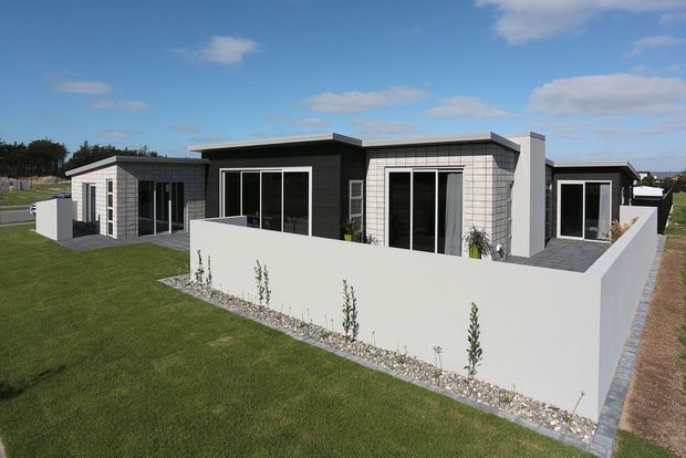 Exterior view of a low-rise contemporary residential development composed of several flat-roofed volumes. The buildings feature white grid-pattern cladding, dark charcoal walls, large black-framed glazed doors, and enclosed private courtyards with white masonry walls. A broad lawn and gravel planting strips surround the property.