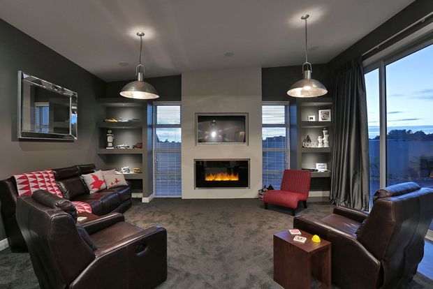 A contemporary living room with dark charcoal walls, built-in shelving, a central fireplace and television, large windows with curtains, pendant lighting, and a leather seating arrangement around a red accent armchair. The room overlooks a blue-toned outdoor landscape through the glazing.