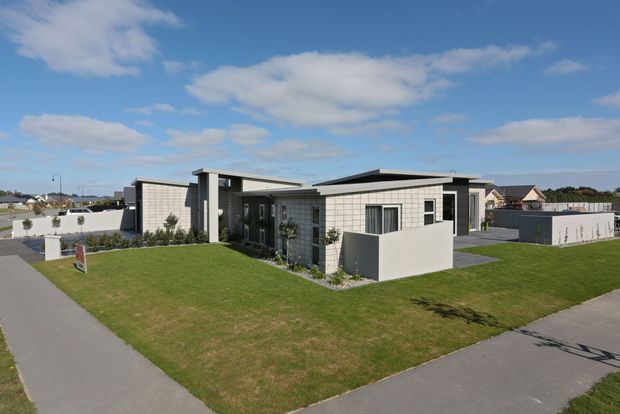 A low, single-storey contemporary home viewed from the street, with a flat and slightly varied roofline, light masonry or blockwork walls, dark window and door frames, and projecting rectangular volumes. The house sits on a landscaped corner site with broad lawns, paved paths, and neighbouring suburban homes visible in the background.