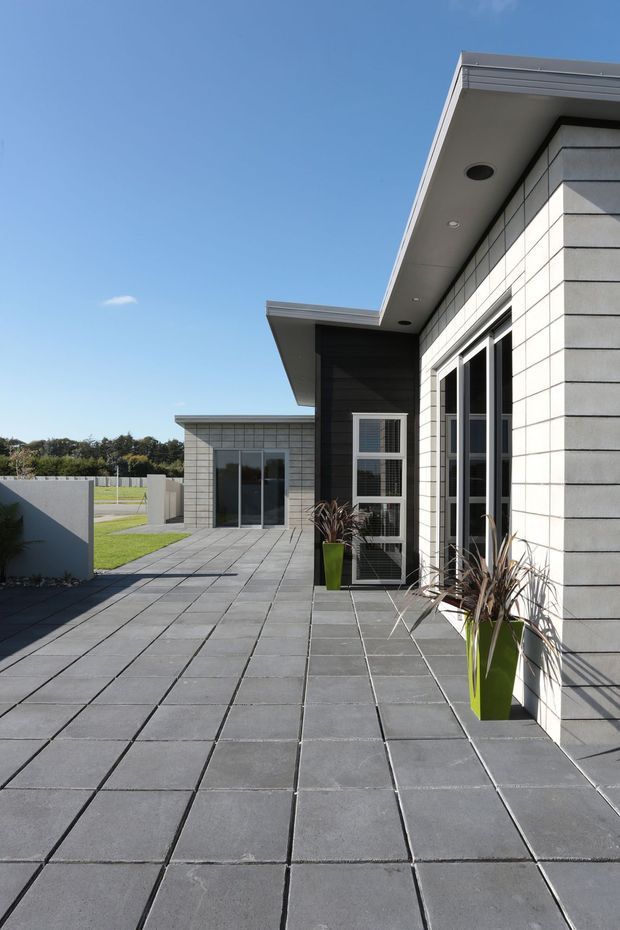 A modern single-storey building exterior with a broad paved terrace or courtyard. The facade combines pale horizontal masonry cladding with dark timber-look wall panels, large glazed doors and windows, and a simple projecting flat roof with recessed downlights. Minimal potted plants line the tiled outdoor area, with a lawn and boundary fencing visible beyond.