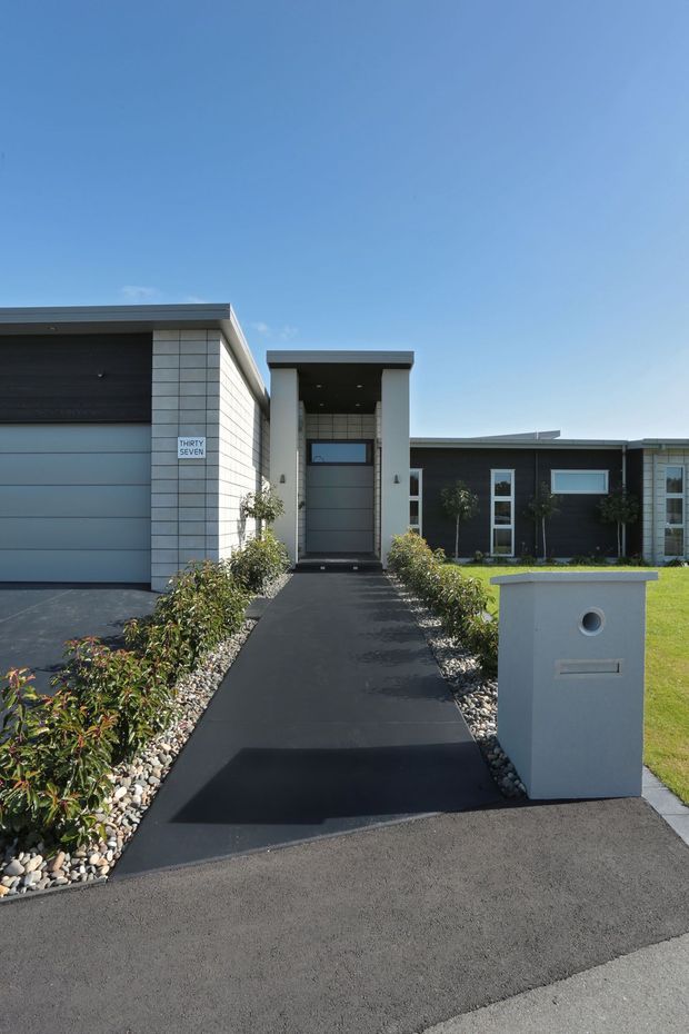 Front exterior view of a contemporary single-storey residence, showing a dark garage and facade, pale masonry cladding, recessed covered entry, landscaped planting beds, and a modern freestanding letterbox beside the driveway.