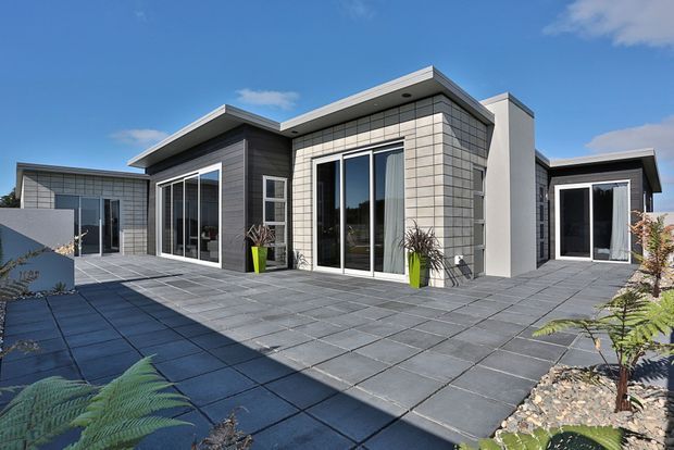 Contemporary single-storey flat-roofed residence viewed from a large paved outdoor terrace. The building has crisp geometric volumes, dark horizontal cladding, light rectangular tile cladding, white rendered sections, and extensive floor-to-ceiling glazed doors and windows. Low-maintenance landscaping with gravel and planting borders surrounds the broad grey paved courtyard.