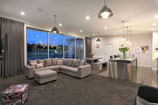 Open-plan contemporary living, dining and kitchen area with a large grey sectional sofa, dark carpet, polished light flooring, extensive glazed doors, a dining table, and a stainless-steel kitchen island. Recessed and pendant lighting creates a bright, modern interior, with curtains framing views to an outdoor area.