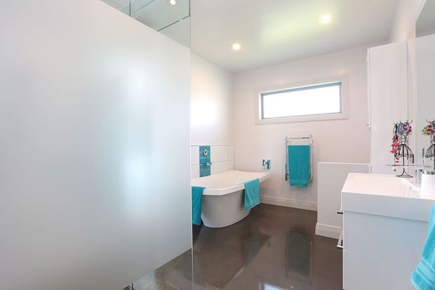 Bright contemporary bathroom with a freestanding white bathtub, white vanity, frameless glass shower enclosure, horizontal window, and glossy dark flooring. Turquoise towels and accessories provide colour accents against pale walls and white fixtures.