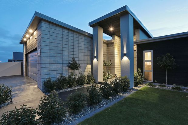 Contemporary single-storey residence viewed from the front at dusk, featuring a dark low-pitched roof, pale horizontally coursed masonry cladding, a projecting entry canopy supported by tall square columns, integrated exterior lighting, a garage, and landscaped garden beds with gravel edging.