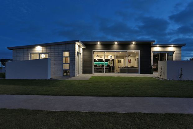 A low, single-storey contemporary home photographed at dusk from the front. The building has a broad flat roof with deep overhangs, dark horizontal cladding at the central entry, light tiled or panelled facade sections, extensive glazed openings, and a brightly illuminated open-plan interior. A neat lawn and paved footpath occupy the foreground.