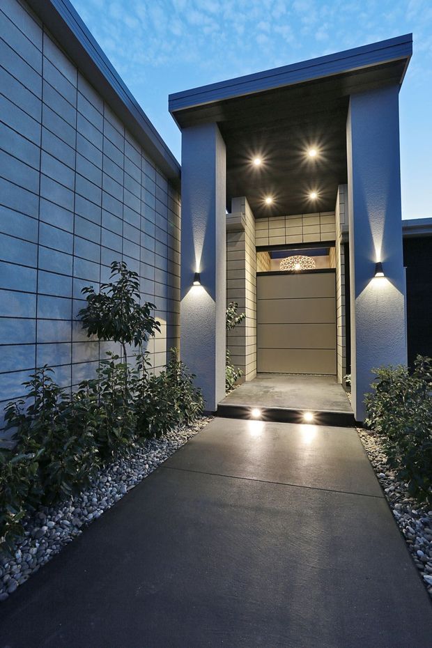 A dramatic contemporary residential entry viewed from an exterior walkway at dusk. A long dark tiled or panelled facade lines the left side, while a tall covered entry portal with broad square columns frames a recessed doorway and illuminated ceiling. Dark concrete paving, integrated wall and step lighting, and narrow planted beds with white decorative stones complete the minimalist approach.