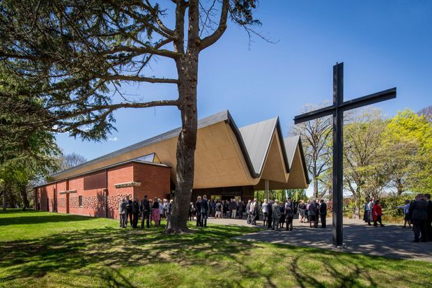 Exterior view of a contemporary church or chapel in a landscaped park setting. The building combines a red brick rectangular wing with a dramatic repeated folded pitched roof forming sharp triangular peaks over the main gathering space. A large black cross stands prominently in the foreground, with people gathered around the entrance and paved walkway. Mature trees and lawns surround the building under a clear blue sky.