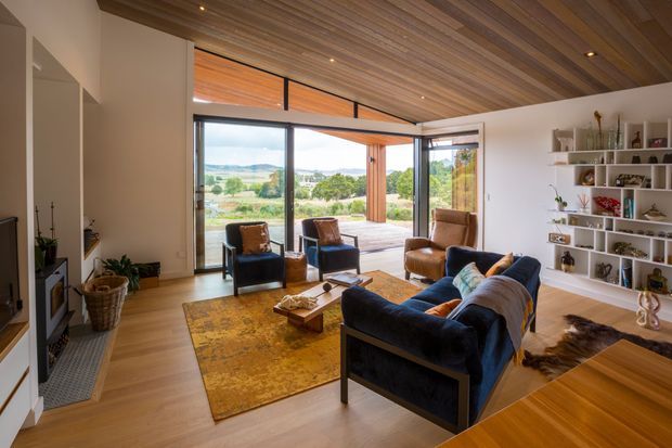 A warm, open-plan living room with floor-to-ceiling glazing opening onto a broad outdoor terrace and landscaped rural view. The room features a timber-lined ceiling, pale timber flooring, white walls, a dark blue sofa, leather lounge chairs, a large patterned rug, a fireplace or media wall, and extensive built-in display shelving. Recessed ceiling lights and generous glazing create a bright, relaxed interior with strong indoor-outdoor connection.