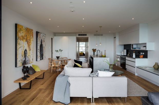 Open-plan contemporary apartment interior combining a living room, dining area and kitchen. A large pale sectional sofa faces away from the camera, with a timber dining table and chairs behind it. The kitchen has white full-height and lower cabinetry, a dark stone-look island benchtop, integrated appliances and pendant lights. Warm timber flooring runs throughout, while white walls and recessed ceiling lights create a bright, clean space. Abstract artworks, plants and small decorative objects add restrained accents.