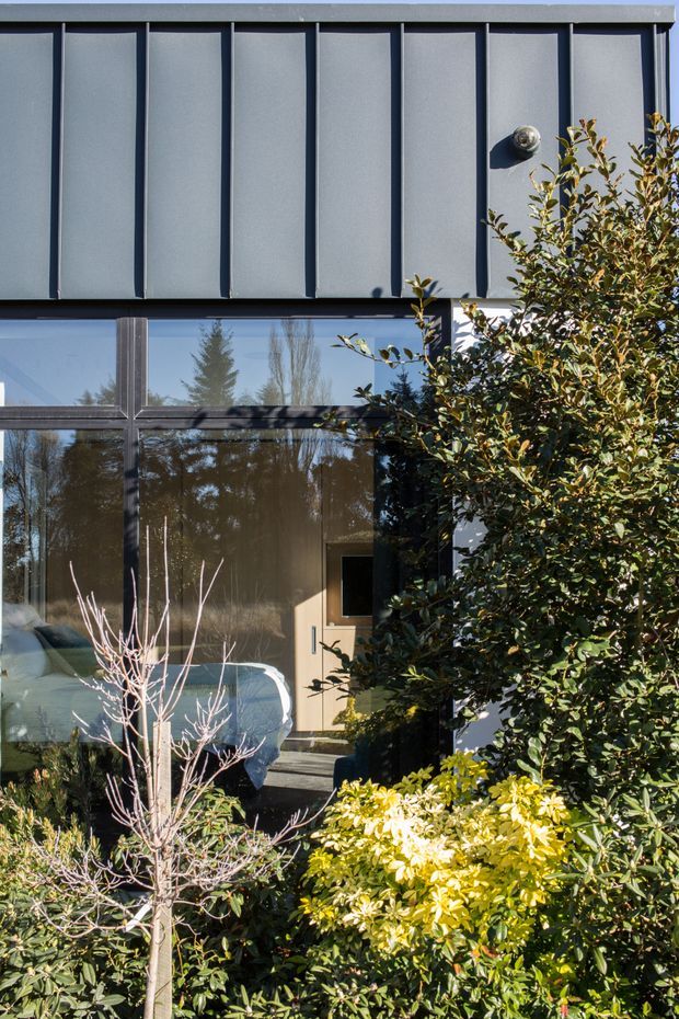Exterior view of a contemporary two-storey facade, partly screened by dense landscaping. The upper level is clad in dark grey vertically ribbed metal panels, while the lower level has large dark-framed glazed openings and a glazed entrance door. Reflections reveal interior furnishings, but the building exterior and planted foreground dominate the image.