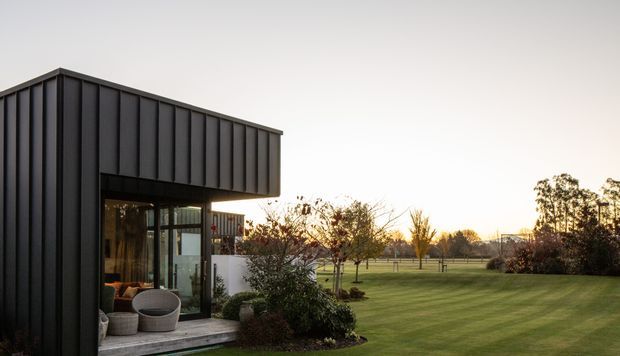 Exterior view of a contemporary single-storey pavilion or extension with dark vertically ribbed metal cladding, a projecting flat-roofed volume, and large glazed openings. The building opens onto a landscaped lawn with young trees and garden planting, creating a strong indoor-outdoor connection.