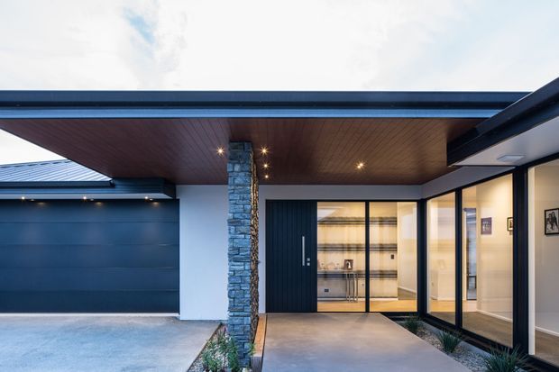 Contemporary single-storey home exterior viewed from the covered entry, featuring dark blue-grey cladding and garage doors, a broad flat roof canopy with warm timber soffit, a stacked-stone column, black-framed glazed entry doors, and a concrete approach. Warm interior lighting is visible through the glazing.