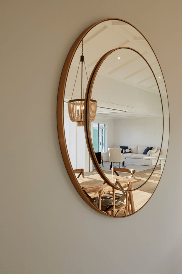 A vertical interior photograph focused on a large oval, warm timber-framed wall mirror. The mirror reflects a bright, refined living and dining area with pale walls, timber flooring, a cream sofa, dining chairs, a timber table, a woven pendant light, and subtle exposed ceiling beams. The composition is calm and elegant, with the mirror serving as the dominant feature.
