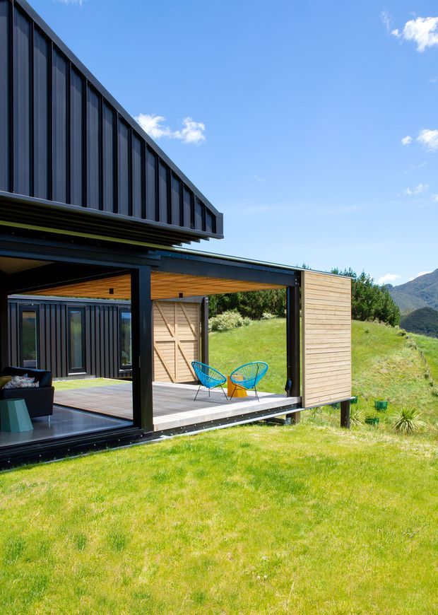 Exterior view of a contemporary pavilion-like house on a grassy sloping site, featuring dark vertical cladding, extensive open glazing, timber-lined soffits, and a covered deck with bright blue outdoor chairs. Mountainous terrain and trees are visible in the background under a clear sky.