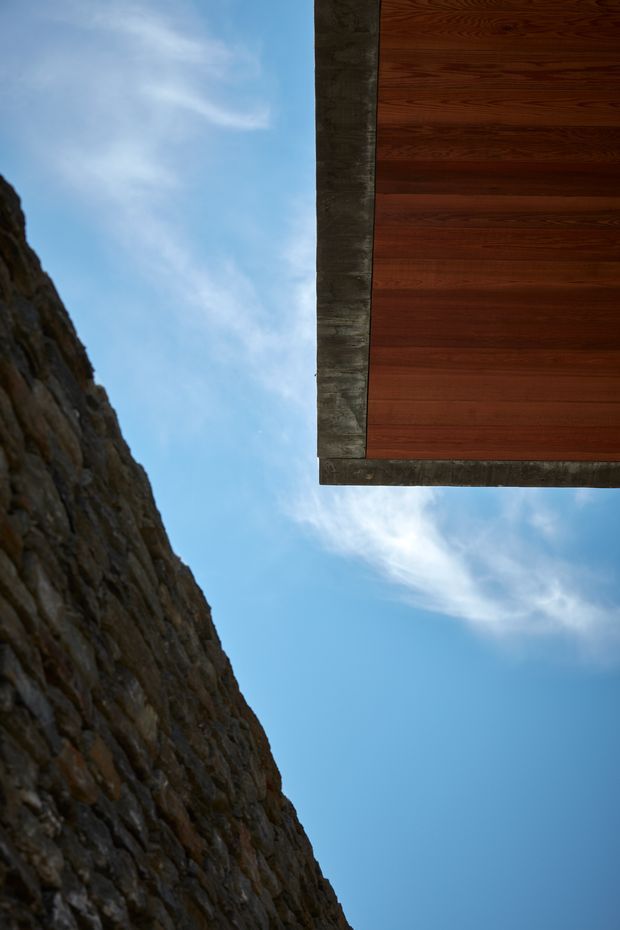 Abstract architectural exterior detail viewed upward between a rugged dark stone wall and the projecting edge of a timber-clad roof or soffit. The image is dominated by blue sky, with strong geometric framing, natural stone texture, weathered concrete edging, and warm reddish-brown horizontal timber boards.