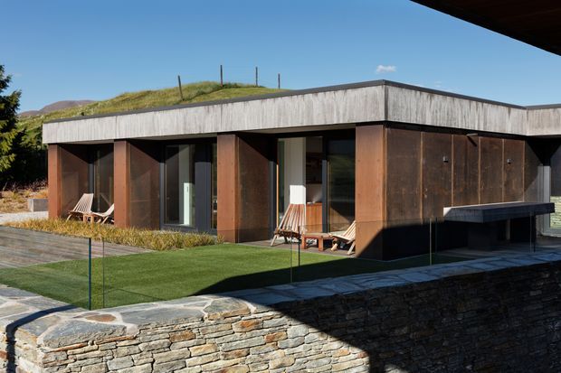 Single-storey modern flat-roof residence set into a landscaped sloping site. The exterior combines dark weathered timber or board cladding with concrete roof-edge bands, extensive glazed openings, a stone retaining wall, paved terrace, and planted green roof. Strong horizontal lines and deep recessed outdoor areas create a restrained contemporary appearance.