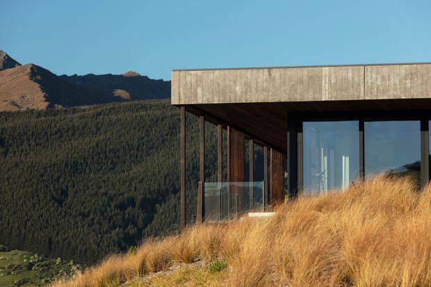 A modern mountain dwelling with a flat roof, weathered timber cladding, dark structural framing, and expansive glazing. The building is partially embedded in a sloping site covered with golden tussock grass, with densely forested mountains visible behind it.