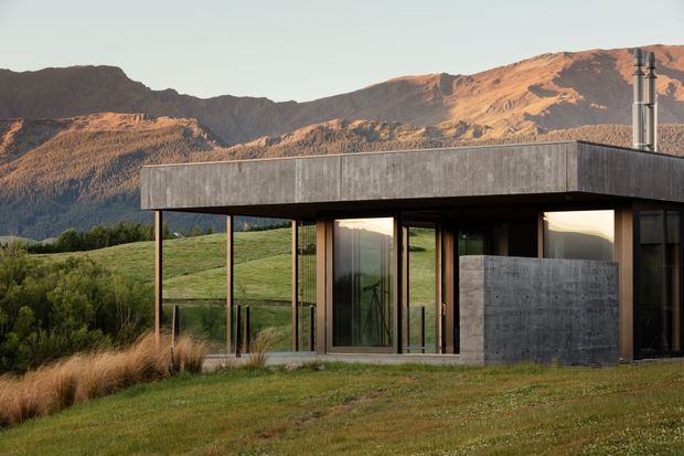 A low, single-storey modern house set in an open grassy landscape with dramatic rugged mountains behind it. The home has a flat roof, dark slender structural posts, extensive floor-to-ceiling glazing, and a mix of weathered grey concrete and dark framing. Warm late-day light highlights the restrained, minimalist composition and strong connection to the surrounding rural landscape.