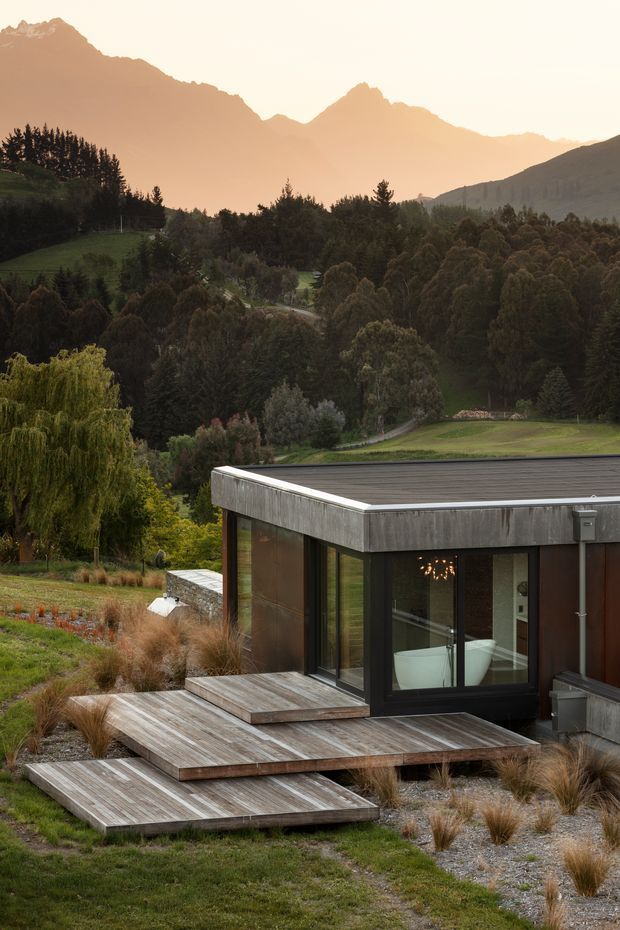 A modern single-storey house set within a landscaped mountain setting. The foreground shows stepped timber decking and gravel paths around a dark-clad pavilion-like building with extensive glazing, a low-profile flat roof, and a visible bathtub and interior lighting through the windows. Rolling lawns, dense trees, and dramatic mountain peaks form the background.