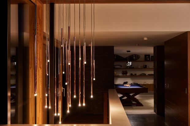 A moody contemporary interior featuring a dramatic cluster of suspended pendant lights with long fine cords and warm exposed bulbs. Timber-framed glazing or wall detailing lines the left side, while dark stone or timber surfaces, open shelving, a billiard table, and dark built-in cabinetry extend into the adjoining space. The composition emphasizes warm lighting, rich wood tones, and deep shadows.
