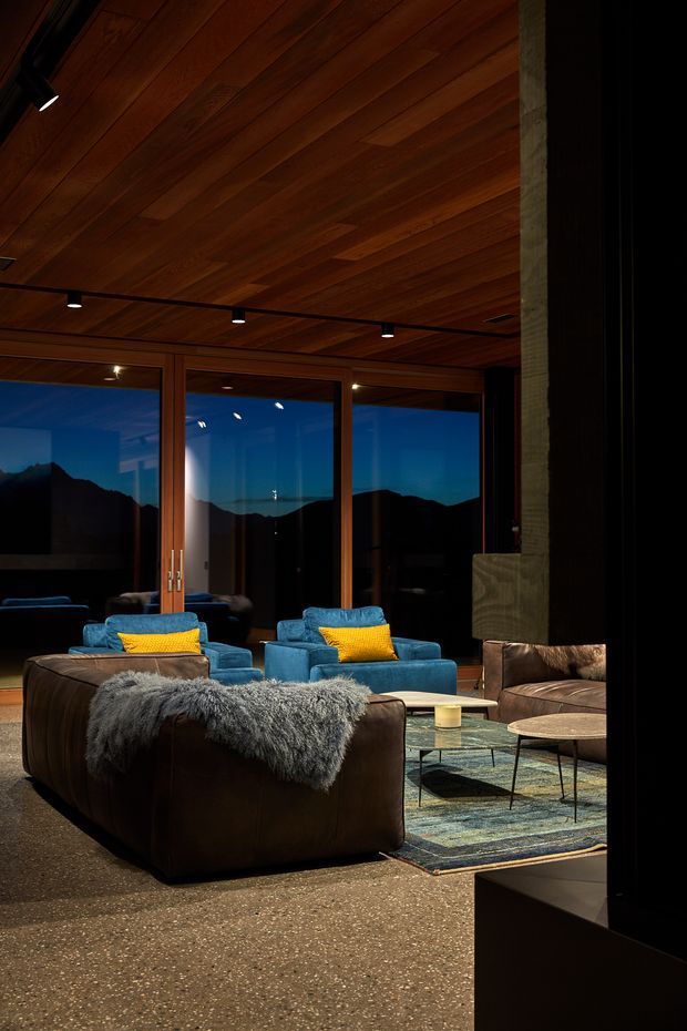 A moody contemporary living room with a warm timber-lined ceiling, dark-framed floor-to-ceiling glazing, and views of silhouetted mountains at dusk. The room contains deep brown leather seating, vivid blue armchairs with yellow cushions, a patterned area rug, low coffee tables, and layered directional lighting. A substantial dark vertical architectural element frames the right side of the image.