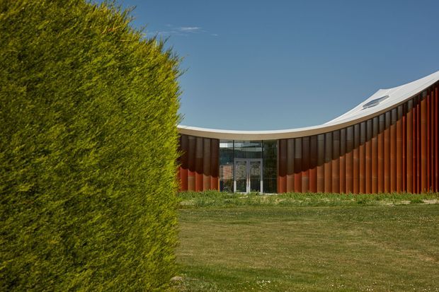 Exterior view of a low, sweeping contemporary building with a pronounced curved roofline and facade. The envelope features closely spaced vertical dark and rust-red cladding elements, with a glazed central entrance and a small roof skylight or clerestory element. A manicured lawn and large clipped hedge occupy the foreground under a clear blue sky.