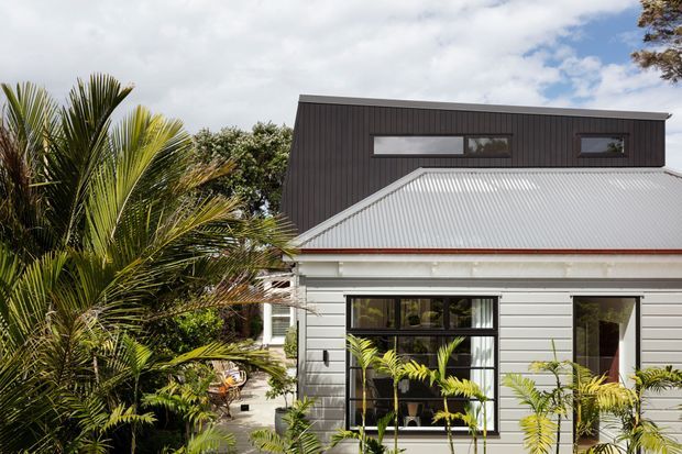 Exterior view of a contemporary residential house surrounded by lush palms and tropical planting. The lower level has white horizontal weatherboard cladding, black-framed glazing, and a light corrugated metal pitched roof, while a dark vertically clad upper-level extension forms a modern box-like volume.