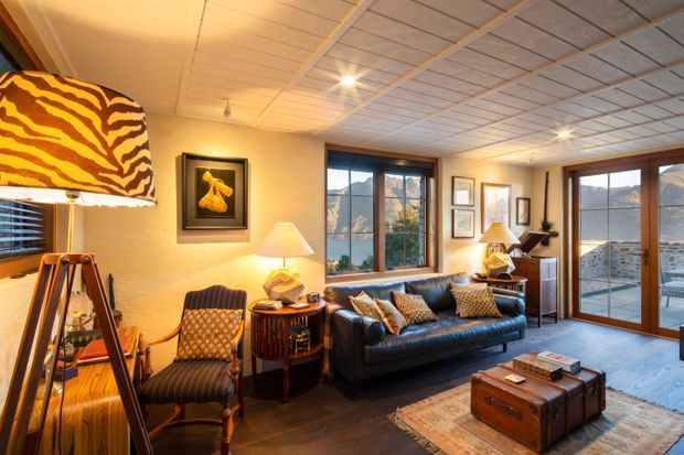 A warm, richly furnished living room with dark timber flooring, a deep blue leather sofa, patterned armchair, vintage-style trunk coffee table, and layered area rugs. Large timber-framed windows and glazed doors provide expansive views toward a mountainous or hilly urban landscape. The room features a sculptural animal-print floor lamp, framed artwork, table lamps, and a suspended acoustic tile ceiling.