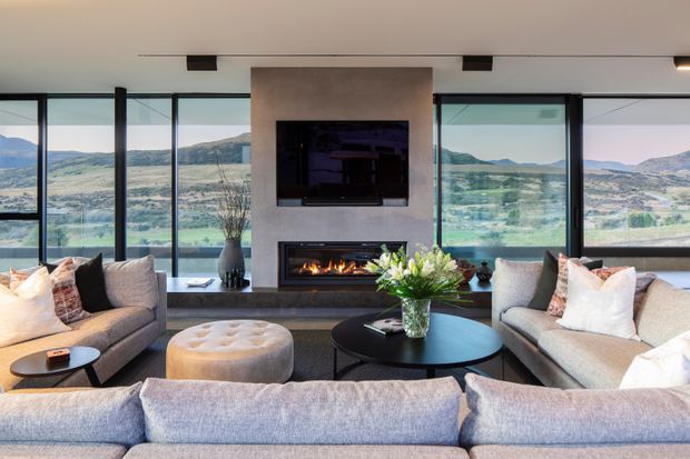 A refined contemporary living room arranged around a central fireplace and television feature wall. Multiple sofas and a round coffee table create a conversational seating area, with expansive black-framed glazing providing panoramic views of a mountainous rural landscape. The neutral upholstered furnishings, pale walls, dark fireplace surround, and warm firelight create a comfortable, polished atmosphere.