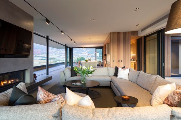 A spacious contemporary living room with a large sectional sofa arranged around nested round coffee tables and a built-in fireplace with television above. Floor-to-ceiling glazing opens the room to expansive mountain and valley views, while warm timber wall panelling, integrated cabinetry, recessed lighting, and a sculptural pendant create a refined lodge-like atmosphere.