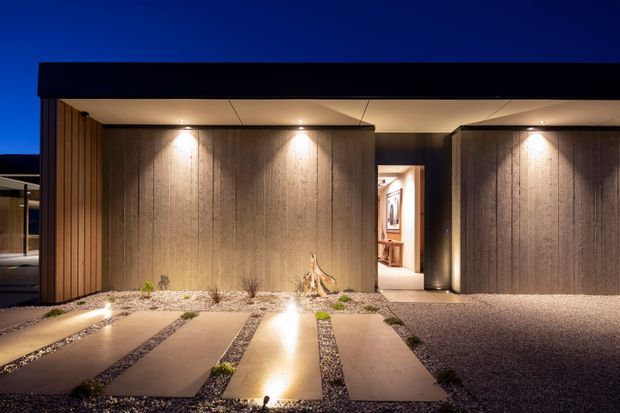 Night-time view of a sophisticated modern house entrance, featuring a strong horizontal flat roof, dark timber or wood-look vertical screening, textured concrete or stone feature walls, recessed warm lighting, and a minimalist gravel forecourt with large rectangular stepping slabs. A warmly lit interior entry is visible through the central opening.