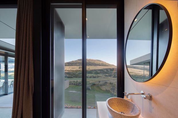 A refined bathroom interior featuring a freestanding textured stone vessel basin on a white vanity, wall-mounted chrome tapware, and a large round black-framed mirror. Full-height dark-framed glazing beside the vanity opens views toward a rural, mountainous landscape, while warm wall lighting and dark curtains create a calm, luxurious atmosphere.
