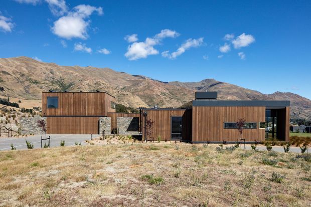 A low-profile contemporary house set in a dry mountainous landscape. The exterior combines warm vertical timber cladding with dark metal roof planes, large glazed openings, and a restrained rectilinear composition. Sparse golden grasses and rocky hills surround the home beneath a bright blue sky.