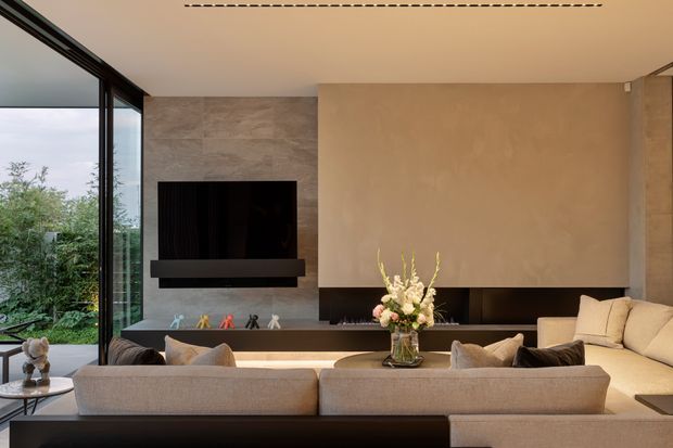 A refined contemporary living room with a large neutral sectional sofa facing a wall-mounted television and long linear fireplace. Full-height black-framed glazing opens views to greenery, while a textured stone-clad television wall contrasts with a warm plaster feature wall. Integrated linear ceiling lighting and restrained furnishings create a calm, luxurious atmosphere.