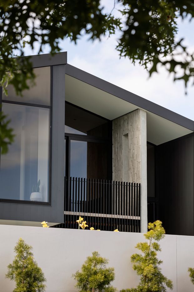 Contemporary multi-level residential facade viewed from outdoors, framed by leafy branches. The building features a strong flat roof canopy, dark vertical cladding, large glazing, a tall textured concrete column, and a black vertical metal balustrade behind a pale rendered boundary wall with landscaped planting.