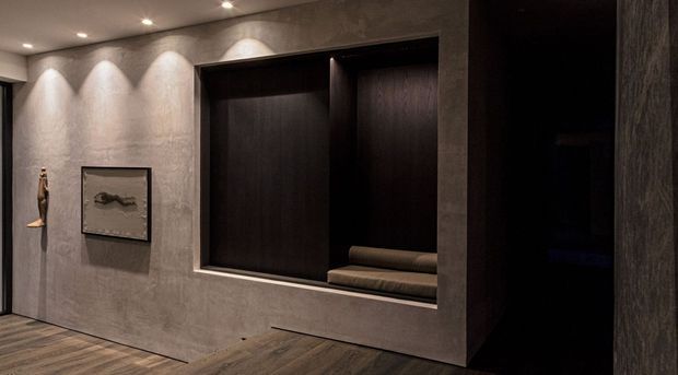 A moody, refined interior featuring a dark built-in recessed display or seating alcove set into a textured grey plaster wall. Integrated ceiling spotlights wash the wall, while dark timber or timber-look joinery, a low upholstered bench, framed artwork, and warm-toned timber flooring create a minimalist, gallery-like atmosphere.