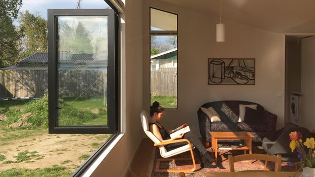 A sunlit contemporary living room viewed from an open glazed doorway or window, with a dark sofa, timber coffee table, armchair, framed artwork, and a person reading. Large black-framed glazing connects the room to a fenced garden and brings in strong natural light. A compact laundry area with a washing machine is visible through the opening at right.