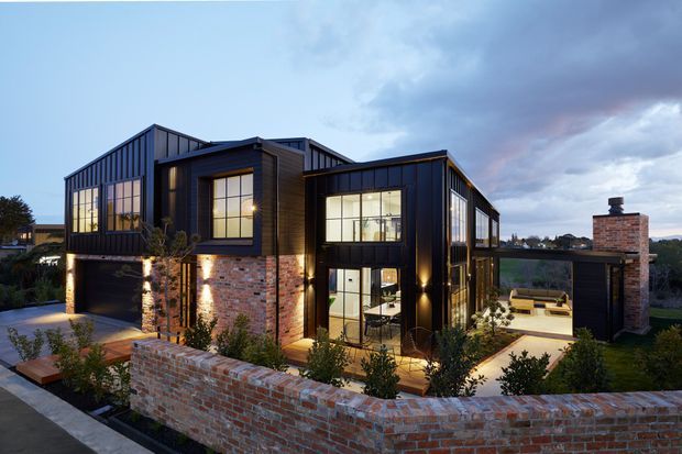 Twilight exterior view of a large contemporary multi-level residence with dark metal and timber cladding, extensive black-framed glazing, exposed red-brick walls, and a covered outdoor entertaining area with a brick chimney. The home sits behind a low brick boundary wall and is warmly illuminated from within and by exterior uplighting.