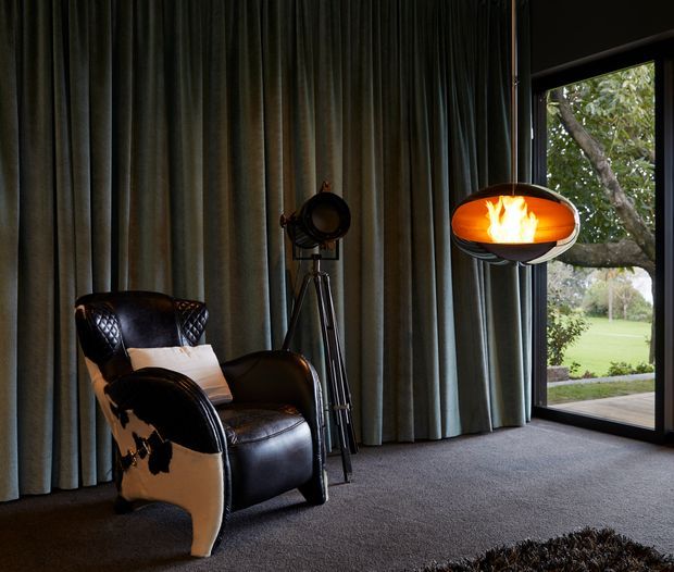 A dark, atmospheric interior lounge or sitting area with a black leather armchair, a vintage tripod floor lamp, a sculptural glowing pendant light, full-height pleated grey curtains, and dark carpet. A large glazed opening provides views to a leafy garden outside.